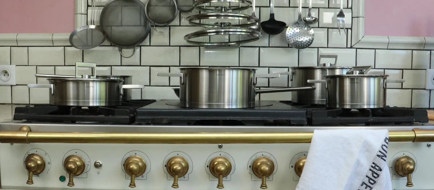 Cristel stainless steel cookware set on a classic French stove with brass knobs, white subway tiles and hanging kitchen utensils
